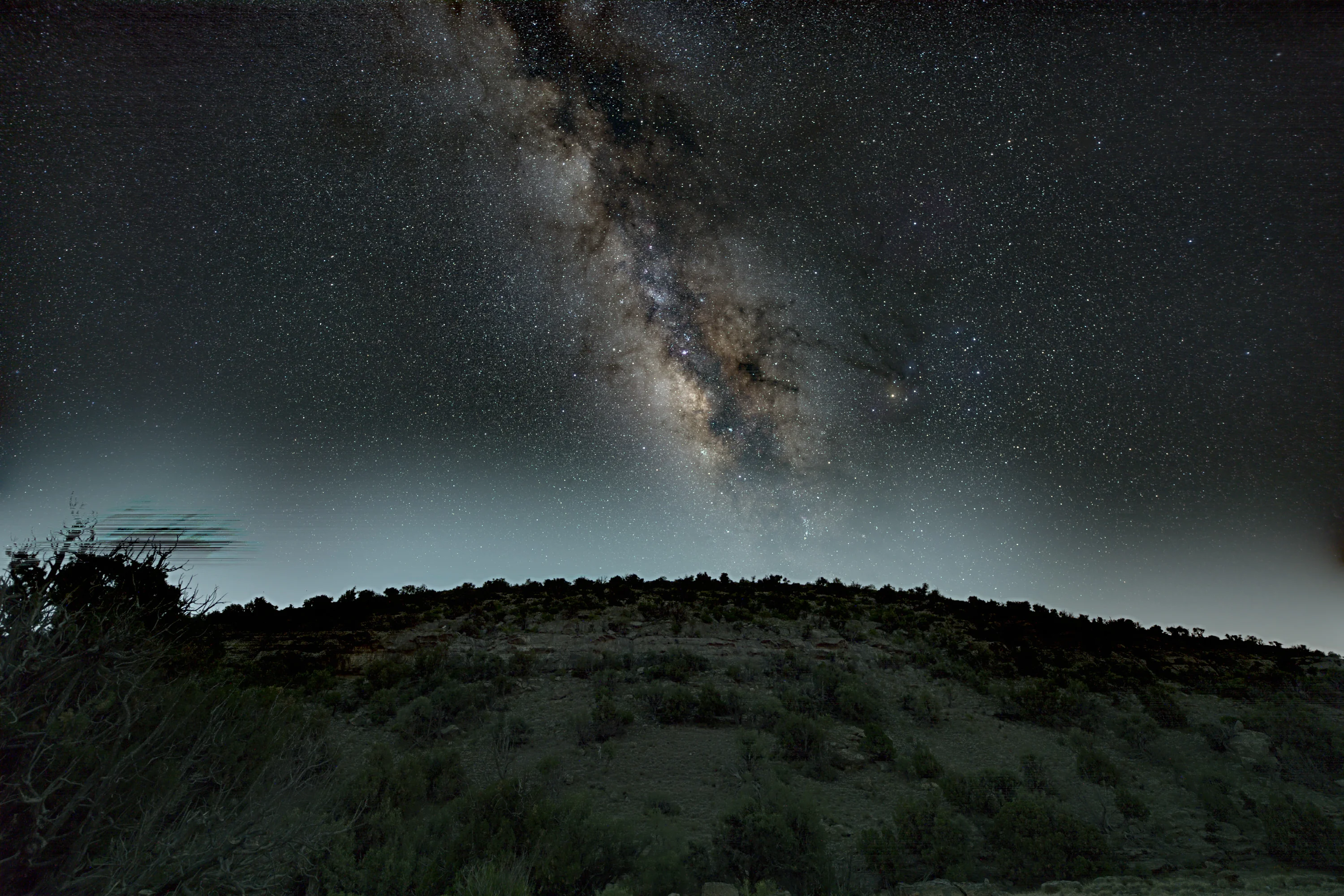 The core of the Milky Way rising above a scrub-covered ridge on Jabal Al Sarah, with a tree silhouetted on the left