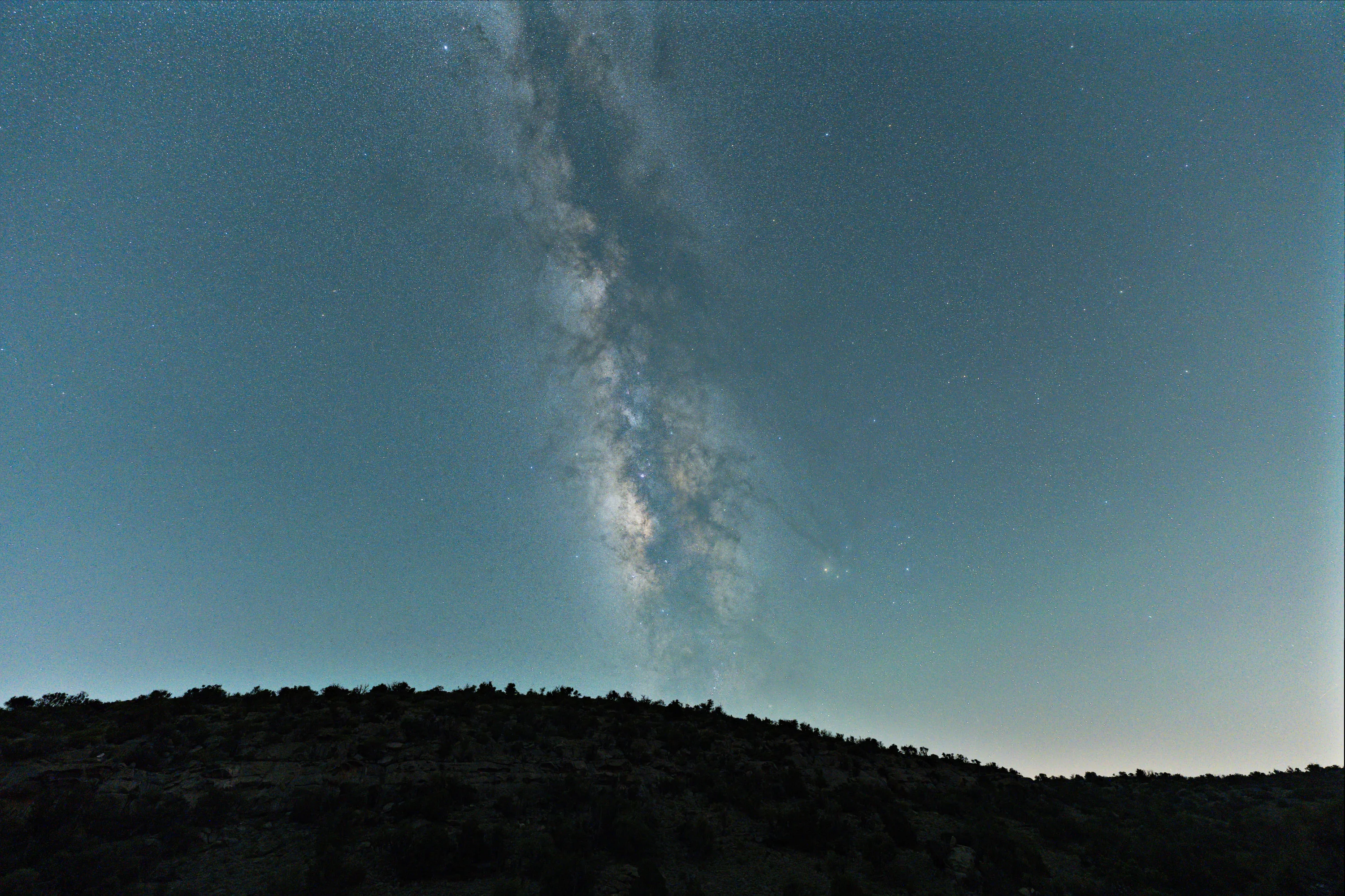 The Milky Way core standing almost upright over the dark line of a scrub-covered ridge, under a blue-green sky